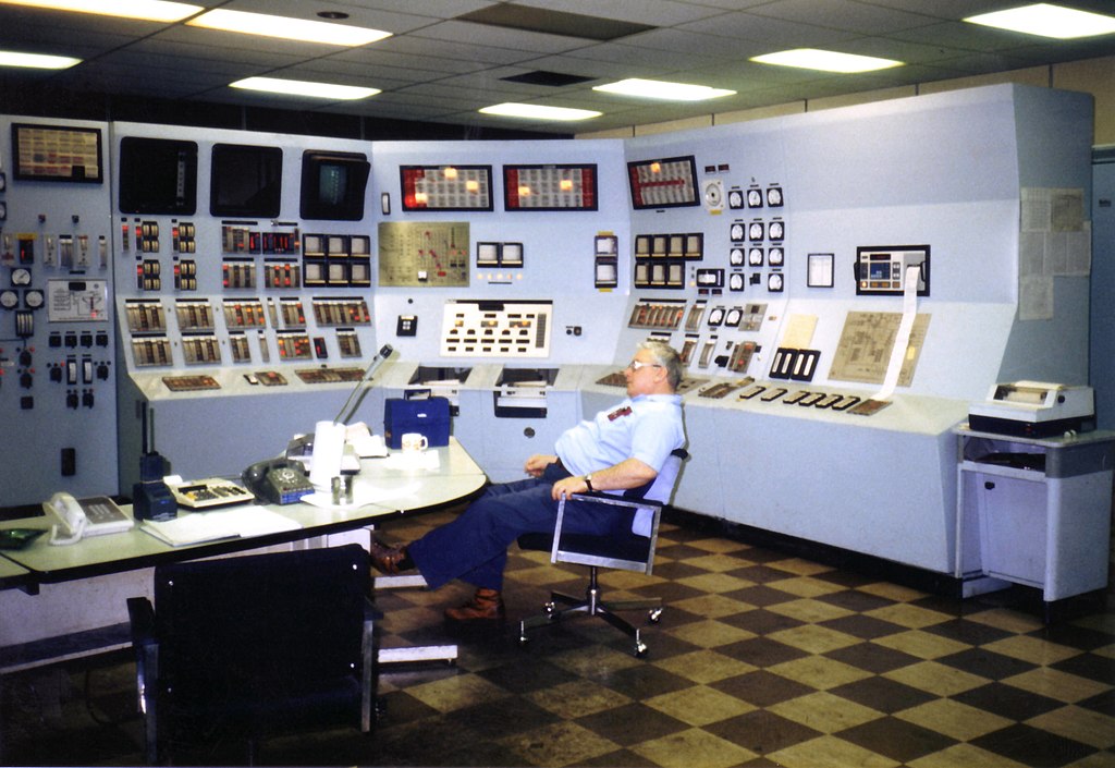 Photo of the interior of the control room in a fossil fuel power plant. The walls at the back are completely covered with large control panels. In front of it an operator sits at a desk in his chair.